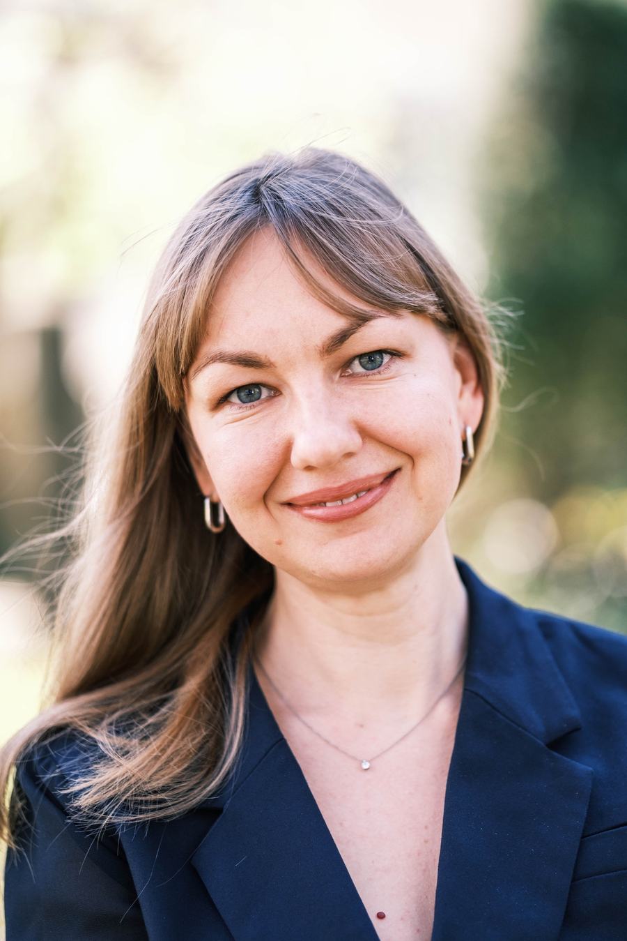 Portrait souriant d’une femme à frange, en blazer bleu marine, dans un jardin ensoleillé
