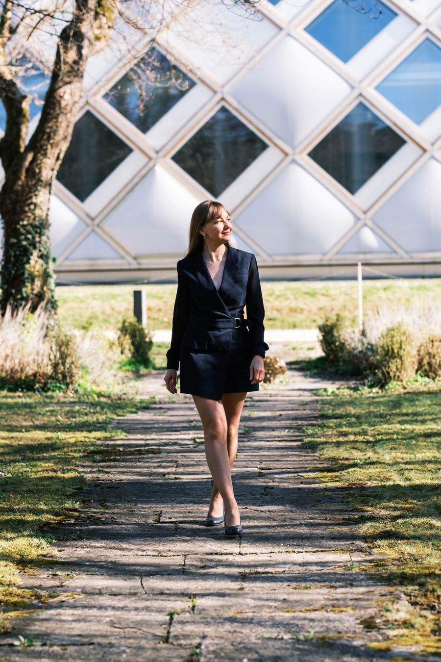 Une femme en tenue sombre marche sur un chemin, devant une façade vitrée en losanges