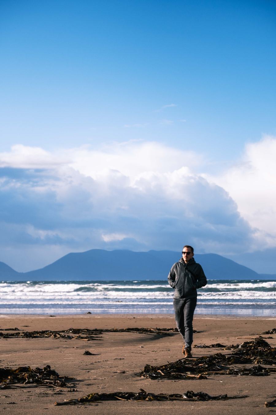 Un homme marche sur une plage, mer et montagnes derrière lui
