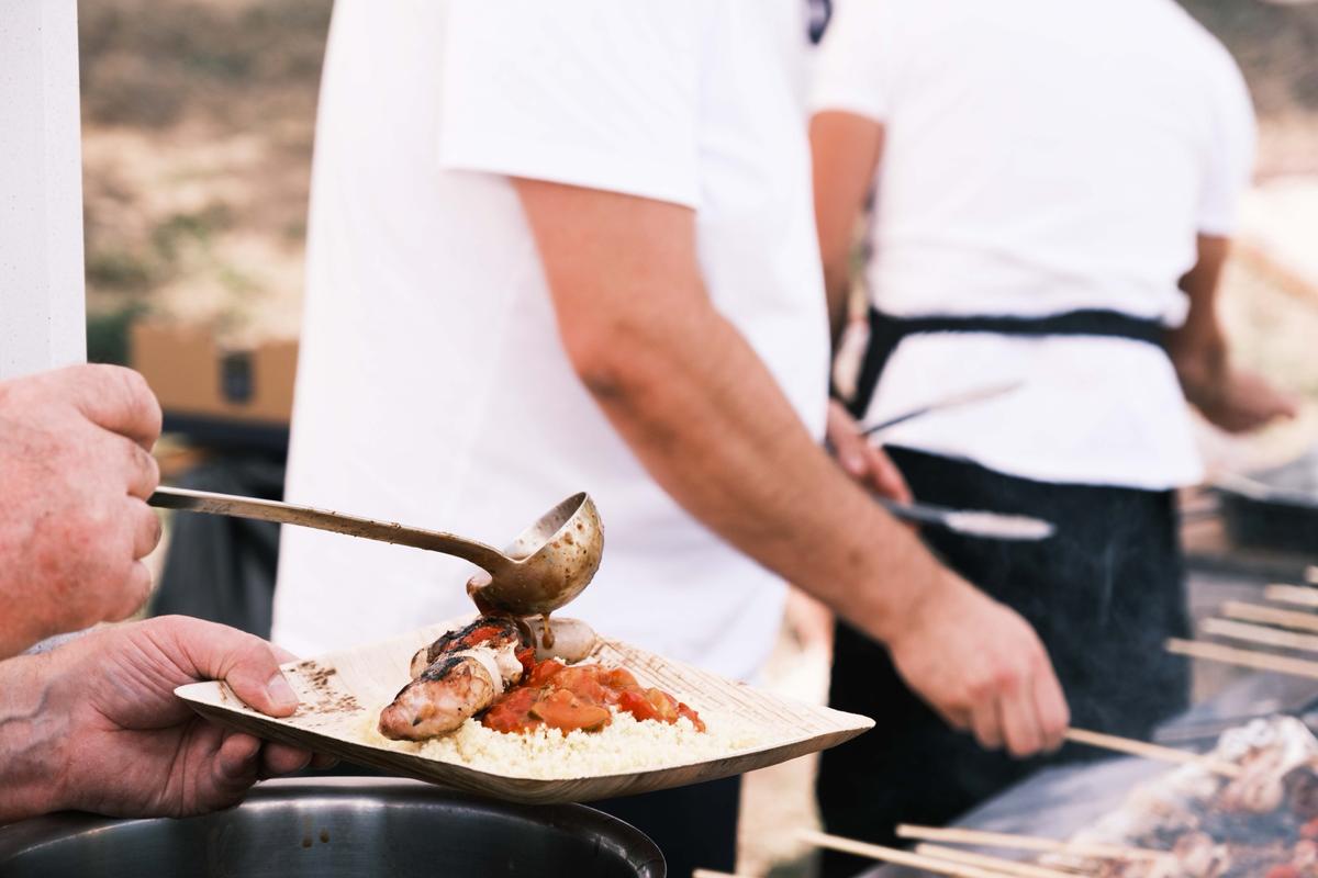 Une louche verse de la sauce sur une assiette de couscous et de brochette, lors d’un repas en plein air
